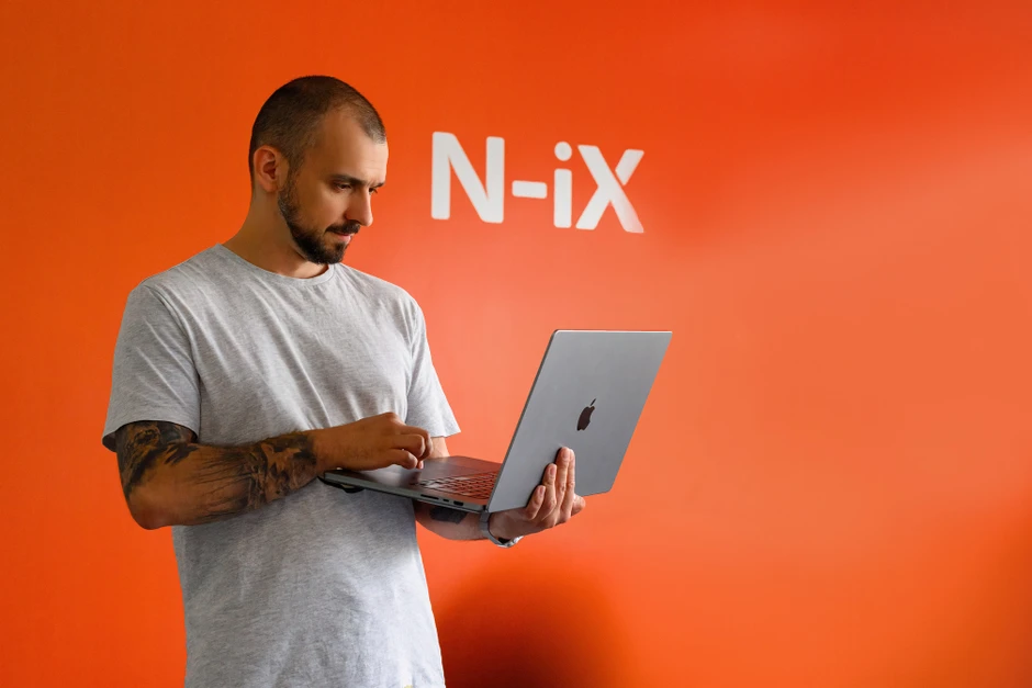 Mężczyzna z tatuażem pracuje na laptopie na tle pomarańczowej ściany z logo N-iX w biurze