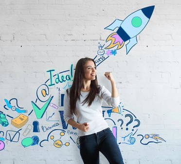 Młoda kobieta z uniesioną pięścią stoi na tle ściany z kolorowymi rysunkami symbolizującymi pomysły i sukces biznesowy
