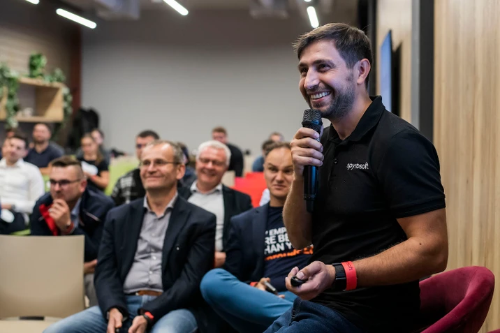 Mężczyzna w koszulce z logo Spyrosoft prowadzi prezentację przed uważną grupą słuchaczy