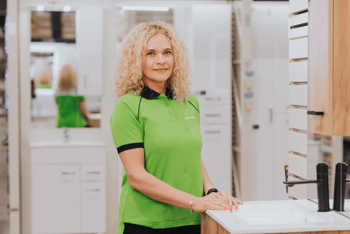 Pracownica w zielonej koszulce Leroy Merlin prezentuje wyposażenie łazienki w sklepie