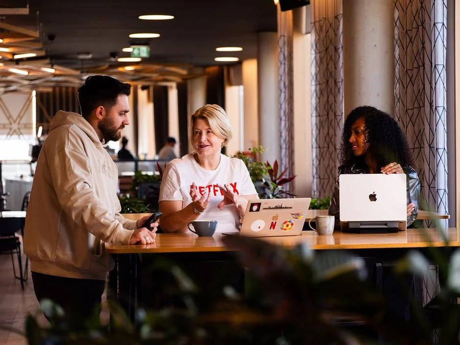 Trzy osoby dyskutują przy wspólnym stole w nowoczesnym biurze z laptopami i filiżankami kawy