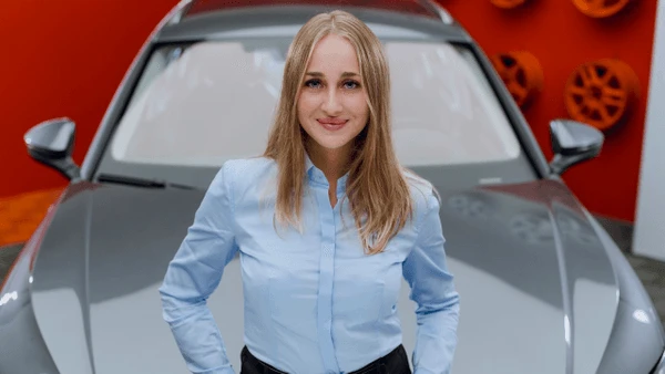 Młoda kobieta w niebieskiej koszuli stoi z uśmiechem przed nowoczesnym samochodem na tle czerwonej ściany