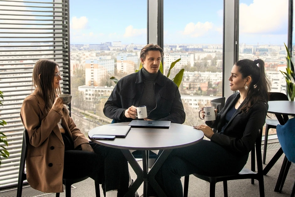 Trzy osoby siedzą przy okrągłym stole w nowoczesnym biurze i rozmawiają przy kawie z kubków firmowych