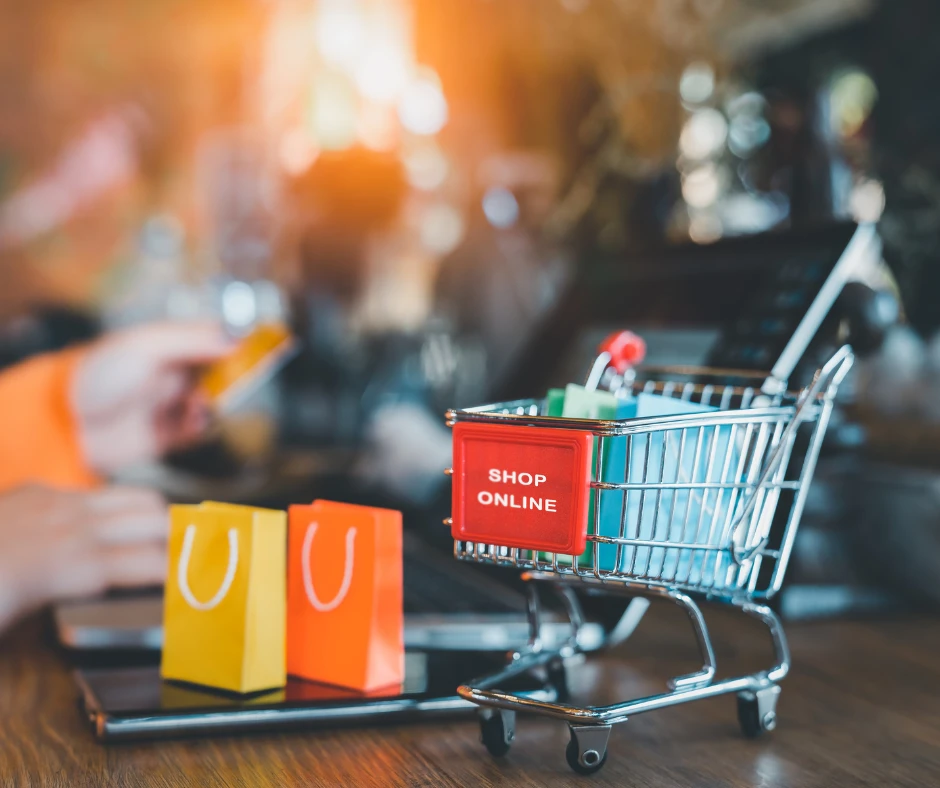 Miniaturowy wózek zakupowy z napisem shop online oraz kolorowe torby na tle osoby płacącej kartą