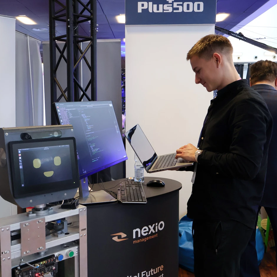 Młody programista pracuje na laptopie przy stanowisku z robotem i monitorem na targach technologicznych Nexio