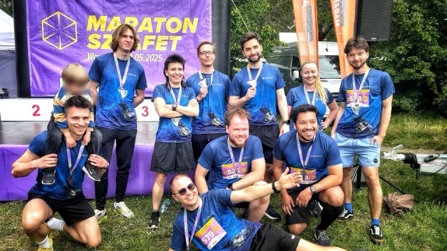 Zespół biegaczy po maratonie sztafetowym w niebieskich koszulkach z medalami na tle baneru eventu