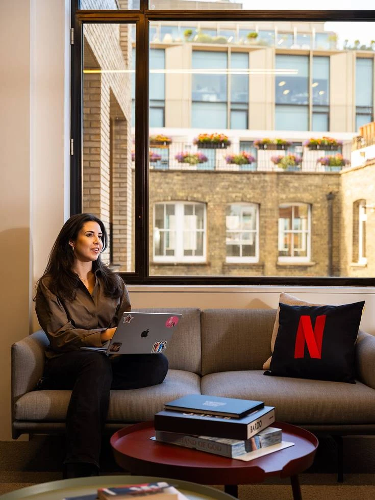 Kobieta pracuje na laptopie siedząc na sofie w nowoczesnym biurze z logiem Netflix na poduszce