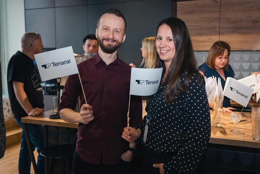 Dwóch pracowników firmy Tenarai uśmiecha się i trzyma firmowe flagi podczas wydarzenia w biurze