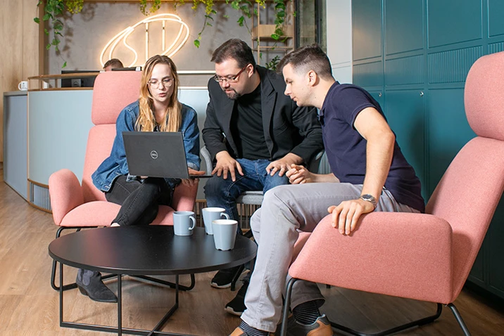 Trzy osoby omawiają projekt przy laptopie w nowoczesnym biurze z wygodnymi różowymi fotelami