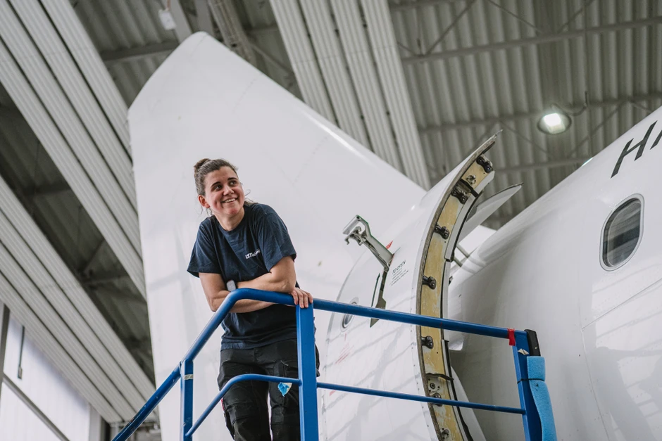 Pracownica w warsztacie lotniczym stoi na platformie przy samolocie i uśmiecha się