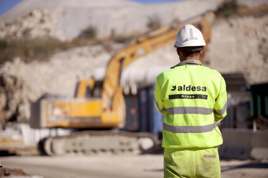 Pracownik budowlany w kasku i odblaskowej kamizelce z logo Aldesa na placu budowy z ciężkim sprzętem