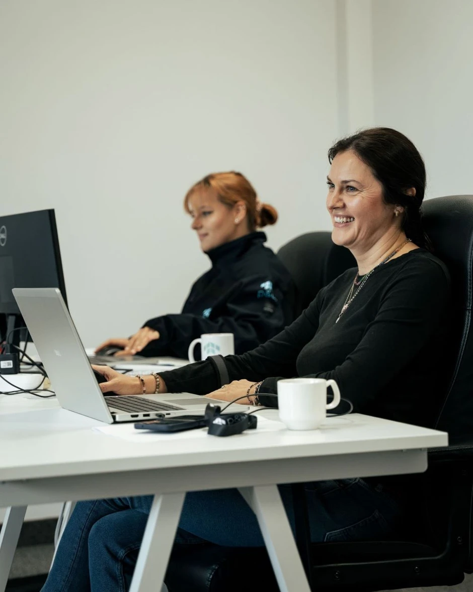 Dwie kobiety pracują w biurze przy komputerach laptop i stacjonarnym, uśmiechnięte i skupione