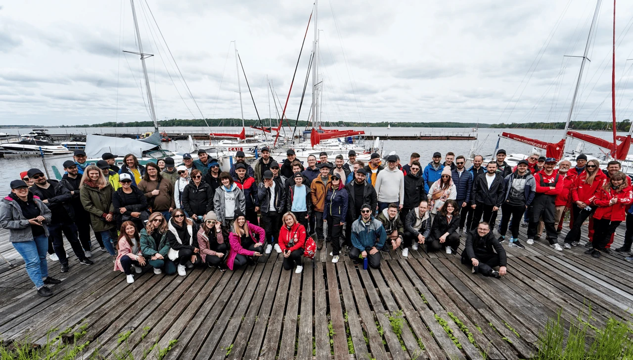 Zespół pracowników na drewnianym pomoście nad jeziorem podczas integracyjnego wydarzenia na łodziach żaglowych