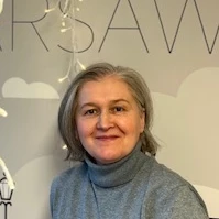 Dojrzała kobieta w szarym golfie uśmiecha się na tle jasnej ściany z napisem w biurowym wnętrzu