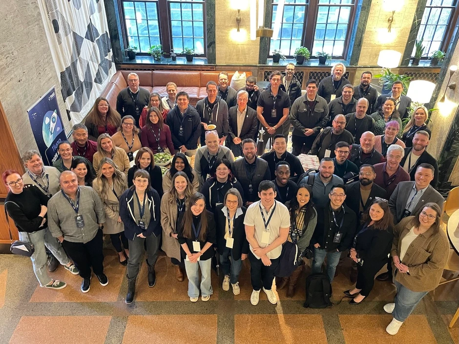 Grupa pracowników pozująca do wspólnego zdjęcia w jasnym pomieszczeniu z dużymi oknami i roślinami