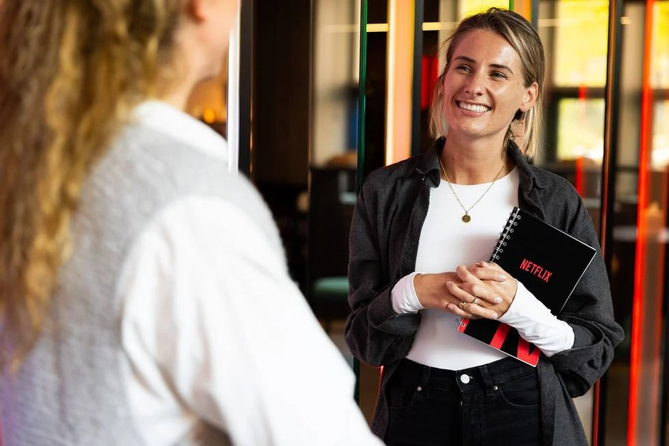 Młoda kobieta z notatnikiem Netflix rozmawia uśmiechnięta w nowoczesnym biurze firmy