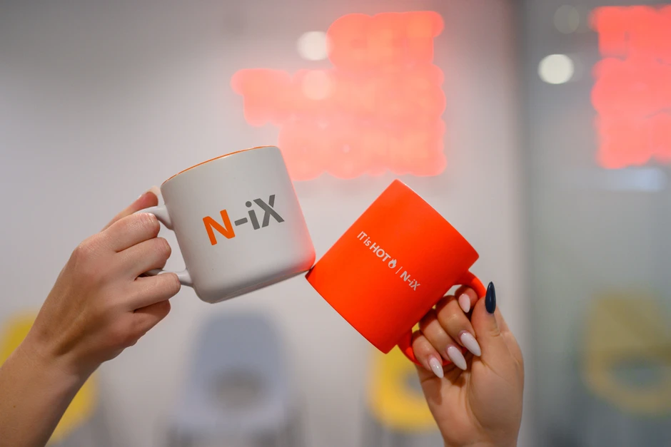 Dwie dłonie stukają się kubkami z logo firmy N-iX w nowoczesnym biurze z rozmytym napisem w tle