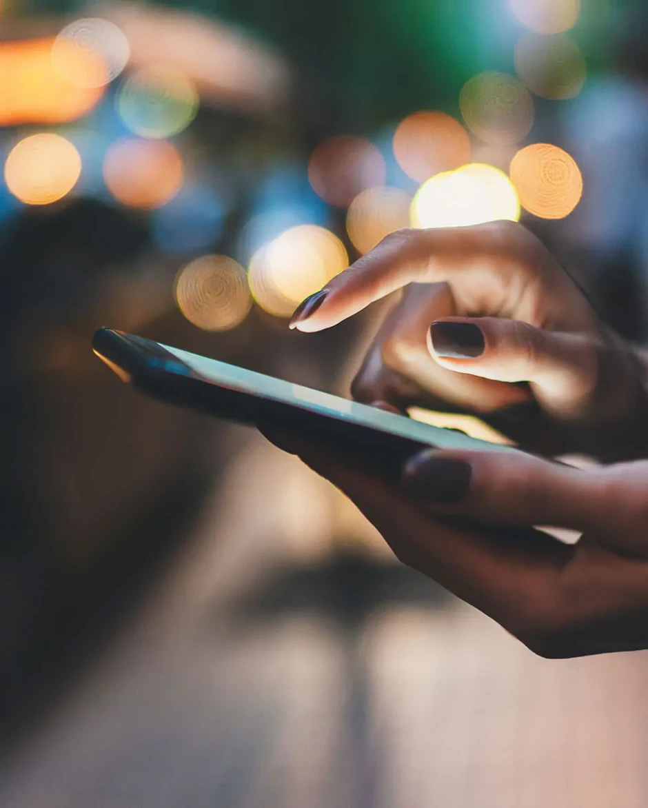 Dłonie z ciemnym lakierem na paznokciach korzystające z smartfona przy rozmytym nocnym oświetleniu ulicznym