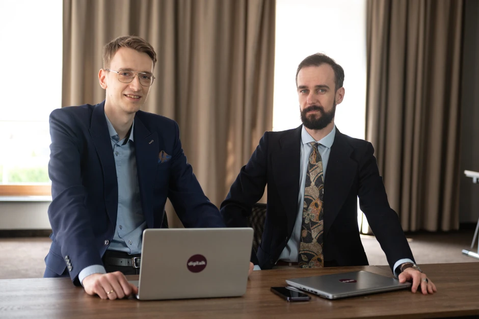 Dwóch mężczyzn w garniturach pracuje przy laptopach w nowoczesnym, jasnym biurze z zasłonami