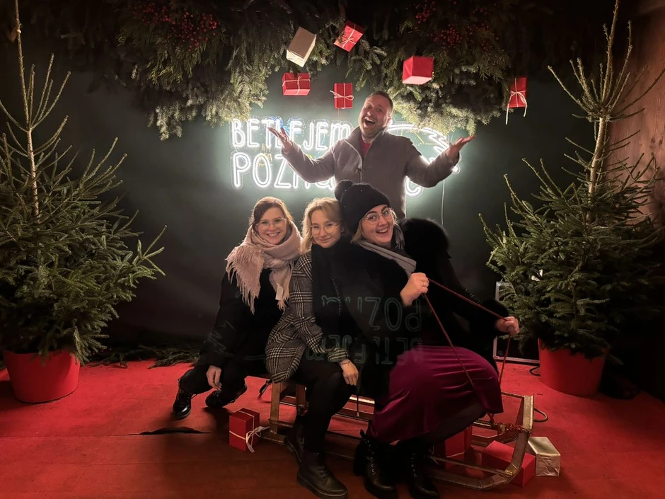 Cztery osoby pozują na sankach w świątecznej dekoracji z choinkami i prezentami
