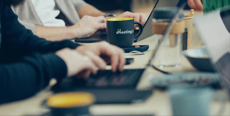 Pracownicy w biurze podczas pracy przy laptopach z kubkiem firmowym dhosting.pl na stole
