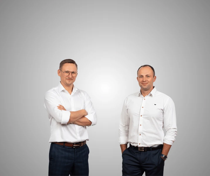 Dwóch mężczyzn w białych koszulach na tle szarej ściany pozujących do firmowego zdjęcia