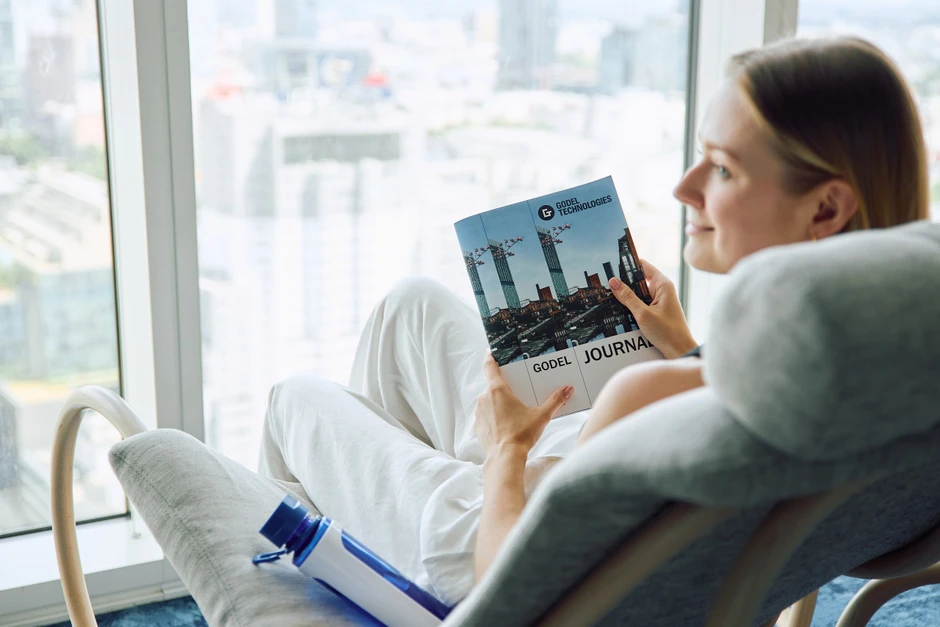 Kobieta relaksująca się w fotelu przy dużym oknie z widokiem na miasto i czytająca magazyn firmowy