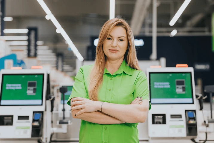 Pracownica sklepu w zielonej koszuli stoi z założonymi rękami przy kasach samoobsługowych