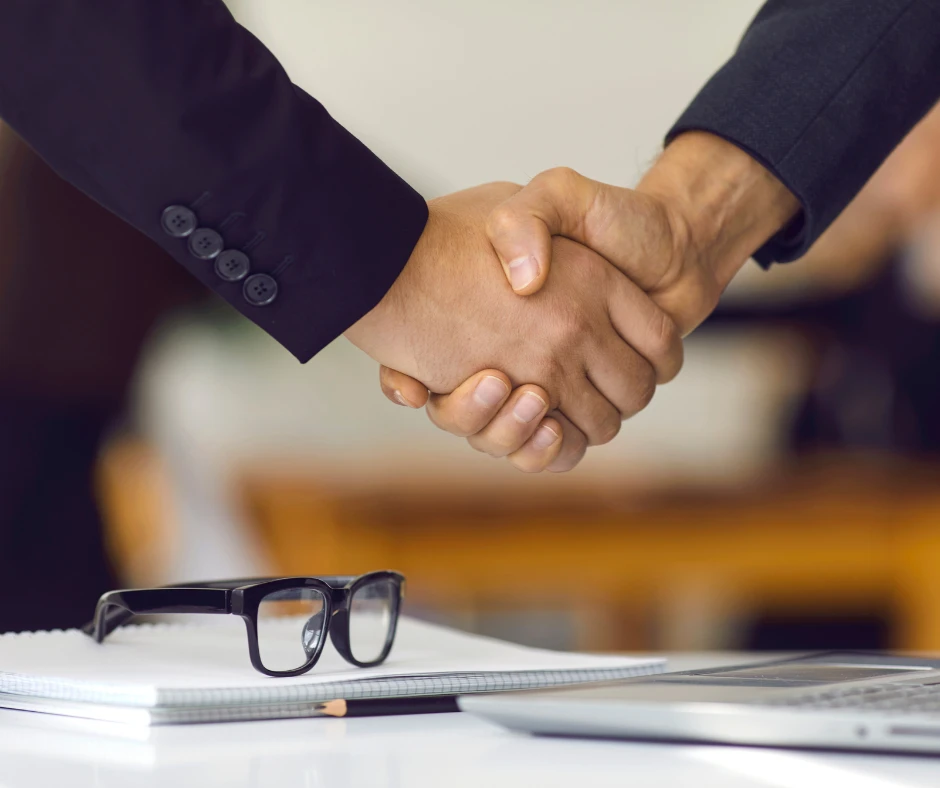 Dłoń dwóch osób w garniturach ściskających się nad biurkiem z notesem okularami i laptopem