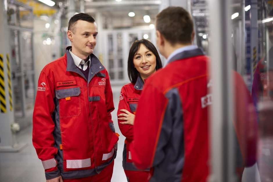 Trójka pracowników w czerwonych mundurach rozmawia w nowoczesnym zakładzie przemysłowym firmy Orlen