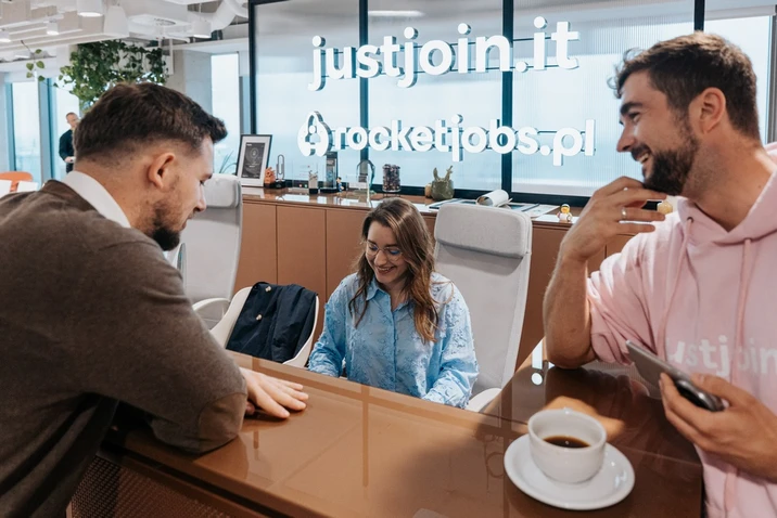 Trzy osoby rozmawiają w nowoczesnym biurze przy recepcji z logo justjoin.it i rocketjobs.pl na szklanej ścianie