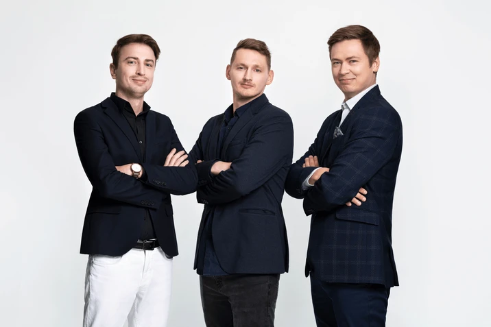 Trzech pewnych siebie mężczyzn w garniturach pozuje z założonymi rękami na jasnym tle