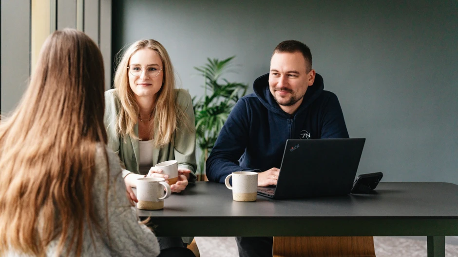 Trzy osoby rozmawiają przy stole biurowym z kubkami i laptopem, w nowoczesnym wnętrzu z rośliną