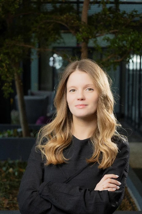 Młoda kobieta z jasnymi włosami w czarnej bluzce stoi z rękami skrzyżowanymi w nowoczesnym biurze