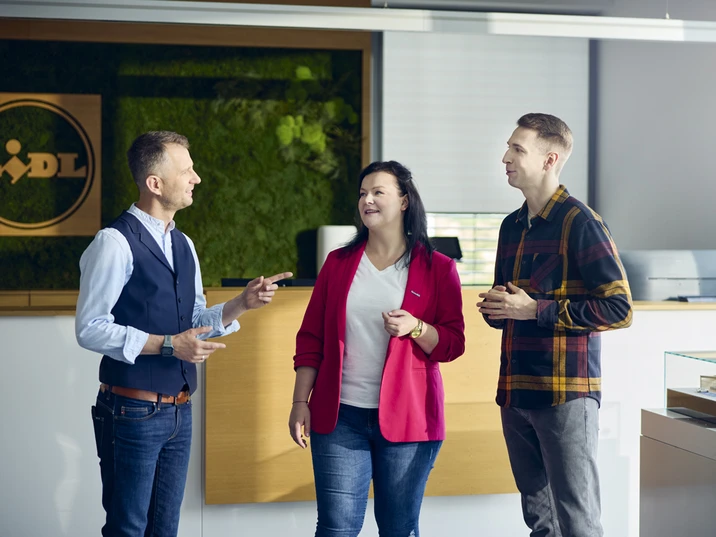 Trzy osoby rozmawiają przy recepcji nowoczesnego biura z zieloną ścianą i logo firmy na tle