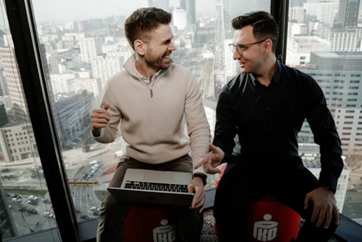 Dwóch mężczyzn rozmawia i uśmiecha się podczas pracy na laptopie w nowoczesnym biurze z widokiem na miasto