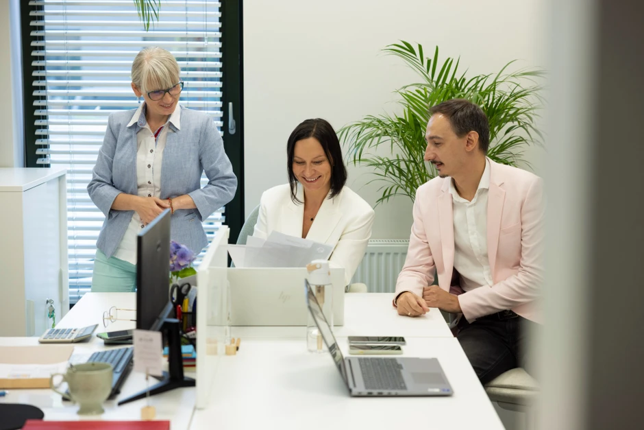 Trzy osoby w biurze omawiają dokumenty przy wspólnym stole z laptopem i komputerem