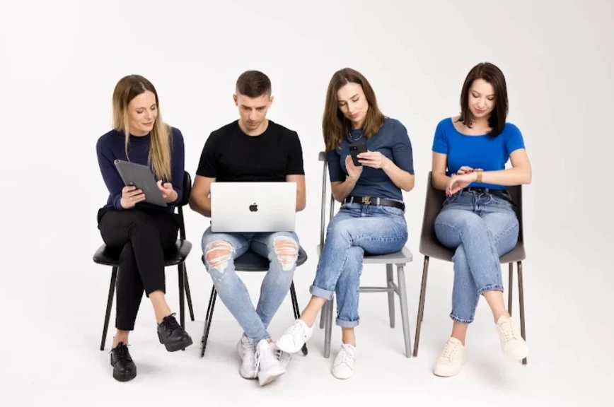 Cztery osoby siedzą na krzesłach, korzystając z laptopa, tabletu i smartfonów w jasnym studio