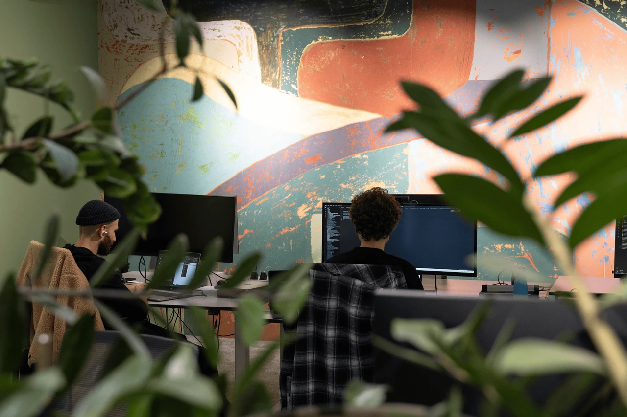 Dwóch pracowników programuje przy komputerach w nowoczesnym biurze z kolorową ścianą i roślinami