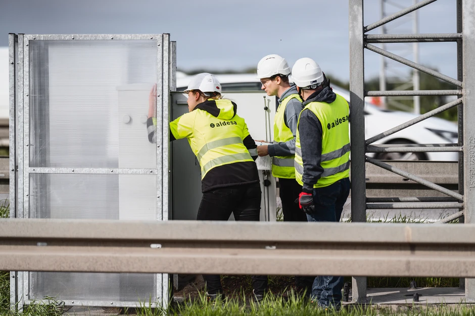 Trzech pracowników w kamizelkach odblaskowych i kaskach zabezpiecza instalację na zewnątrz przy drodze