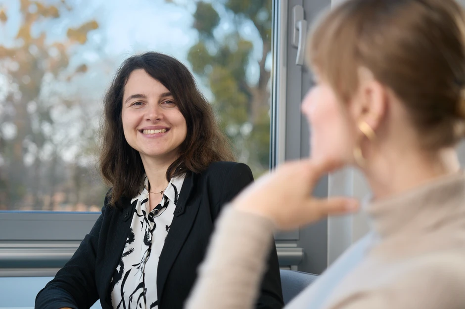 Uśmiechnięta kobieta w biurze rozmawia z koleżanką na tle okna z widokiem na drzewa i niebo