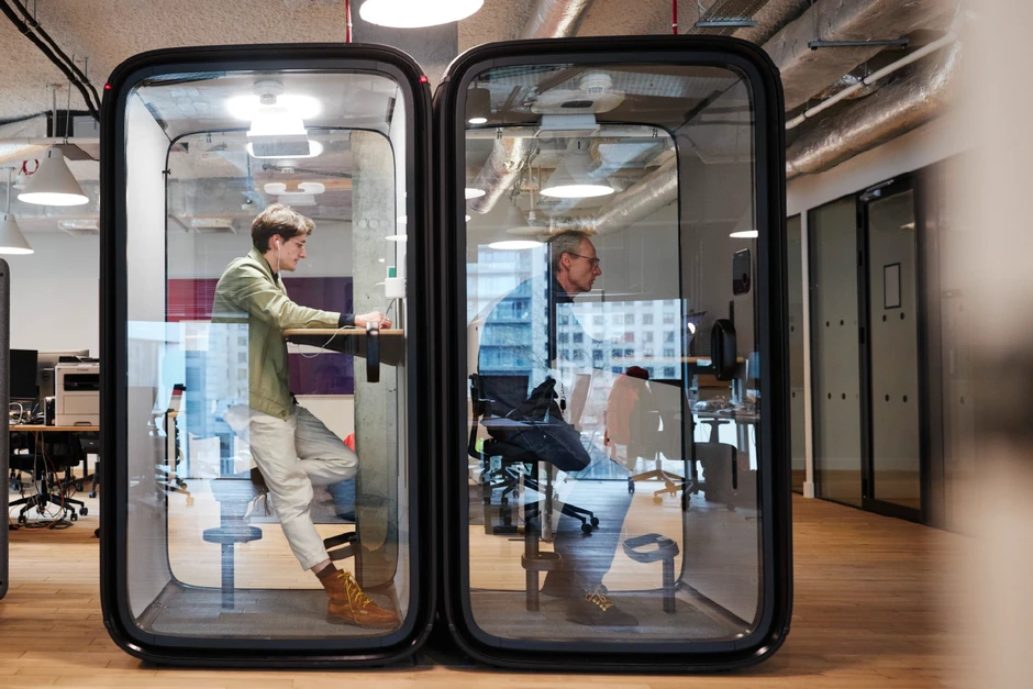 Dwóch pracowników korzysta z nowoczesnych, przeszklonych kabin do pracy w otwartym biurze z drewnianą podłogą