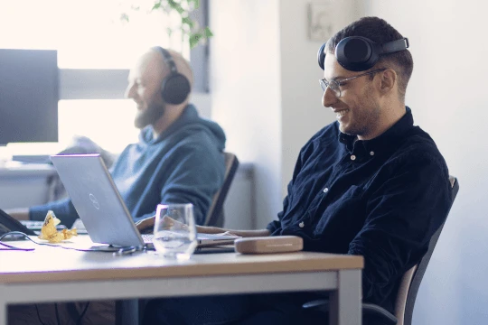 Dwóch mężczyzn pracuje w biurze, korzystając z laptopów i słuchawek, atmosfera jest przyjazna