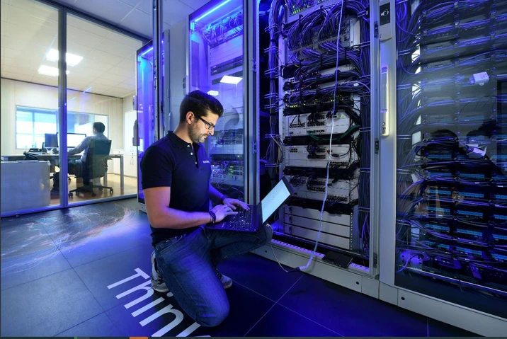 Technik IT pracuje przy serwerach w nowoczesnej serwerowni o niebieskim oświetleniu biurowym
