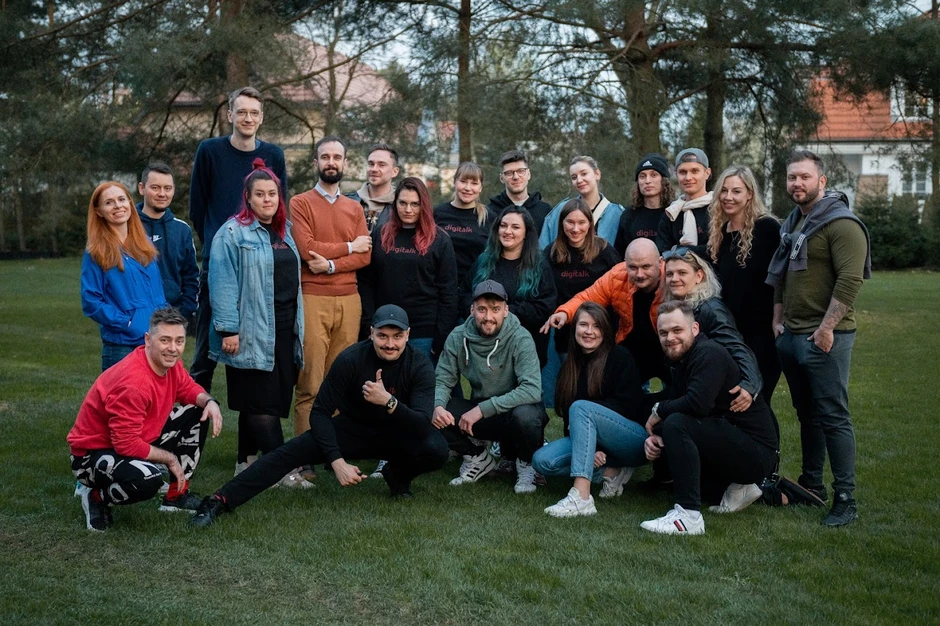 Zespół firmy pozuje na zewnątrz na trawie w otoczeniu drzew i budynków w tle