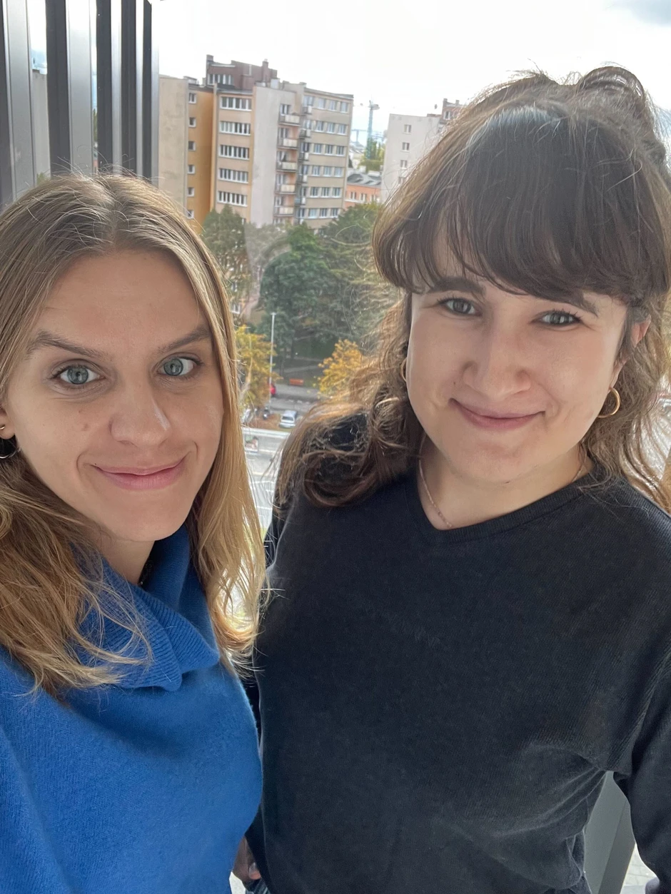 Dwie uśmiechnięte kobiety pozują w jasnym biurze z widokiem na miejskie budynki za oknem