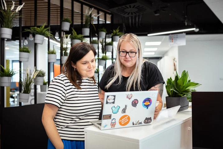 Dwie kobiety wspólnie pracują przy laptopie w jasnym biurze z roślinami i nowoczesnym wystrojem