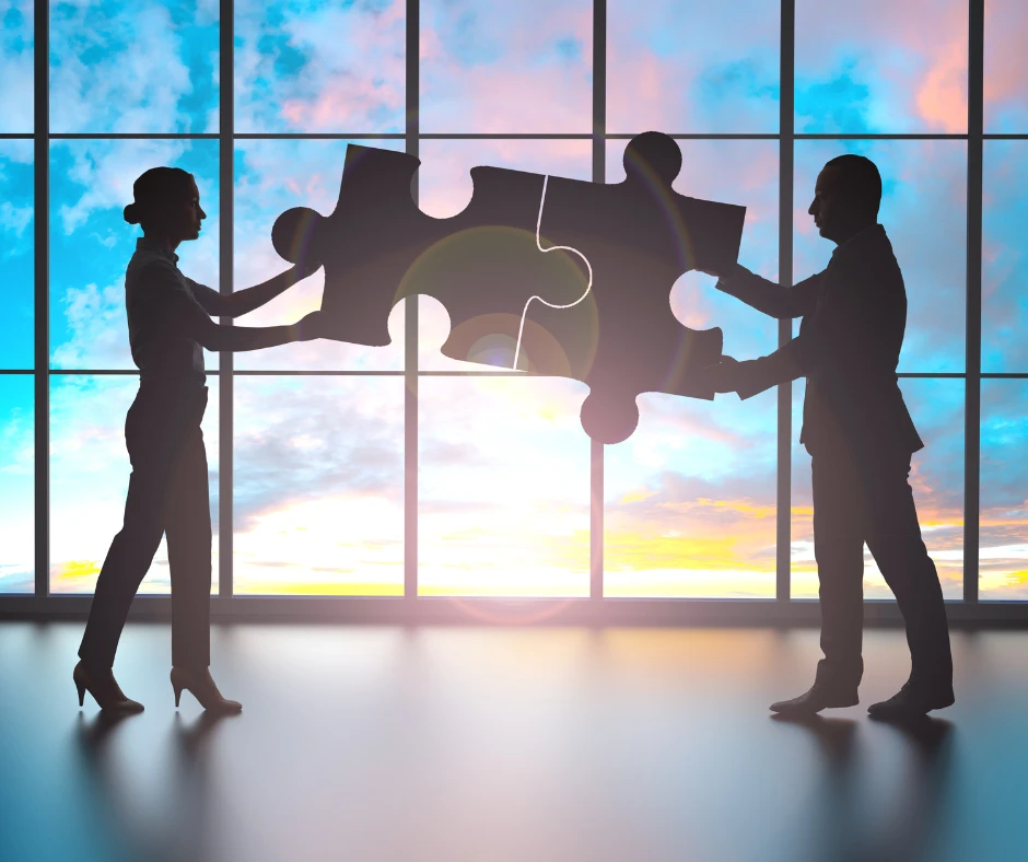 Dwóch pracowników łączy puzzle na tle dużych okien z widokiem na niebo o zachodzie słońca symbolizując współpracę