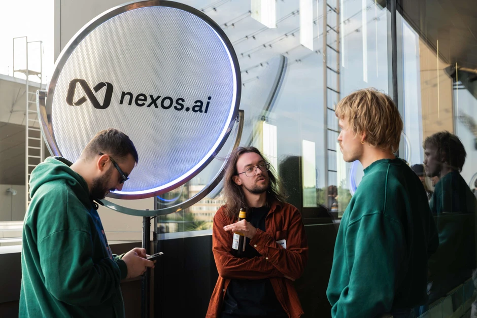 Trzech mężczyzn rozmawia przed podświetlanym logo nexos.ai podczas firmowego spotkania na tarasie