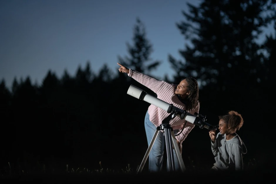 Kobieta i dziewczynka oglądają gwiazdy przez teleskop pod nocnym niebem w otoczeniu drzew